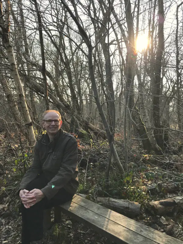 Donnie sitting on a bench in a woodland. The trees are bare of leaves and the winter sun is a yellow ball showing between the trees. Donnie is a white male with short red hair and is wearing brown framed glasses.