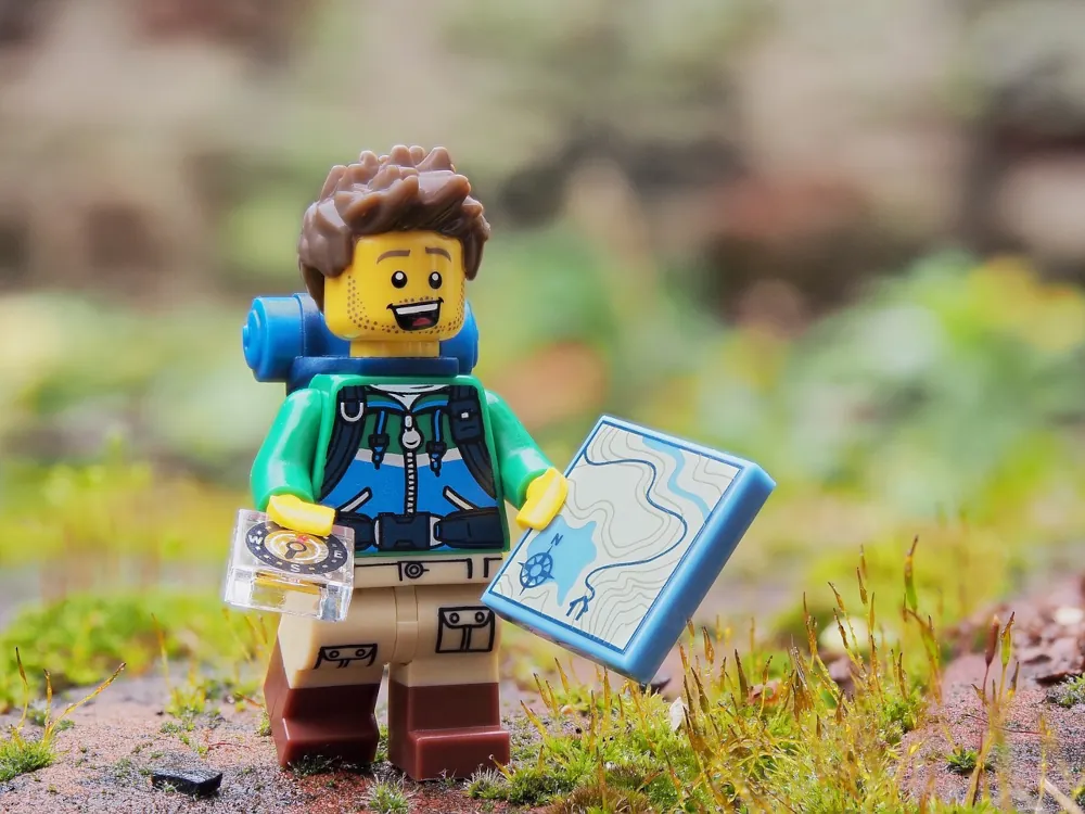 A Lego figure of a man hiker. He is wearing brown boots, beige cargo pants and green top with blue panels. He has a rucksack on his back and is carrying a map in one hand and a compass in the other. He has short brown hair and has a stubble beard. He looks very happy as he walks in a real grassy and rocky environment.