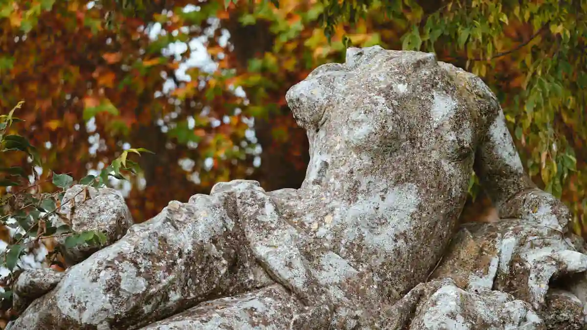 There is a backdrop of trees with the leaves turning autumn colours. In the foreground on carved stone is a statue of a reclining woman in the classical style. She is missing her head and arms. The whole statue is grey and weathered with lichen patches.